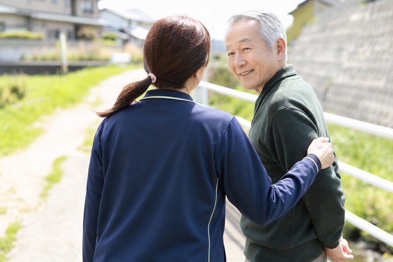 晴れた日の散歩道で、スタッフと腕を組んで笑顔で歩く利用者さま